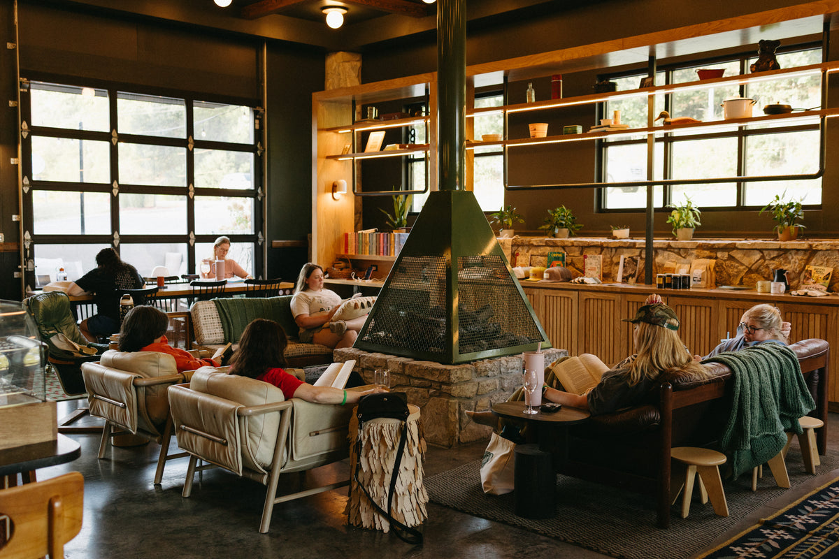 A cozy indoor reading lounge with several people sitting and reading around a central stone fireplace. The space has large industrial-style windows, wooden shelving, and comfortable seating including armchairs and sofas.